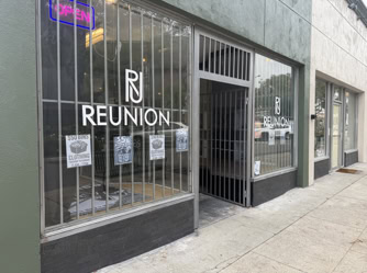 Storefront of Reunion clothing shop with barred windows, open sign, and $5 clothing bin posters displayed in the front windows.