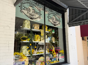 A storefront window for 2nd Ave Boutique featuring a vibrant, all-yellow display. Multiple shelves are packed with lemon-patterned backpacks, handbags, artificial sunflowers, and summer-themed decor. The shop is housed in a white brick building with a grey metal awning, and a street scene i